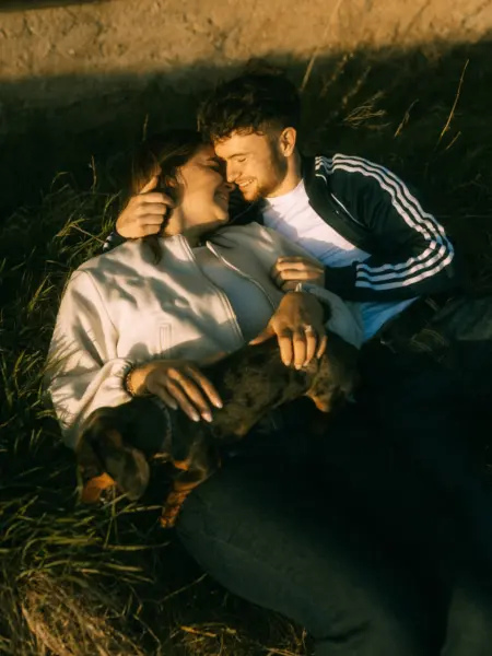 Ein verliebtes Paar kuschelt bei goldenem Sonnenuntergang im Gras, während sie ihren kleinen Dackel halten. Die intime Szene fängt eine echte Lovestory ein – perfekt für authentische Pärchenfotos in Schwerin.