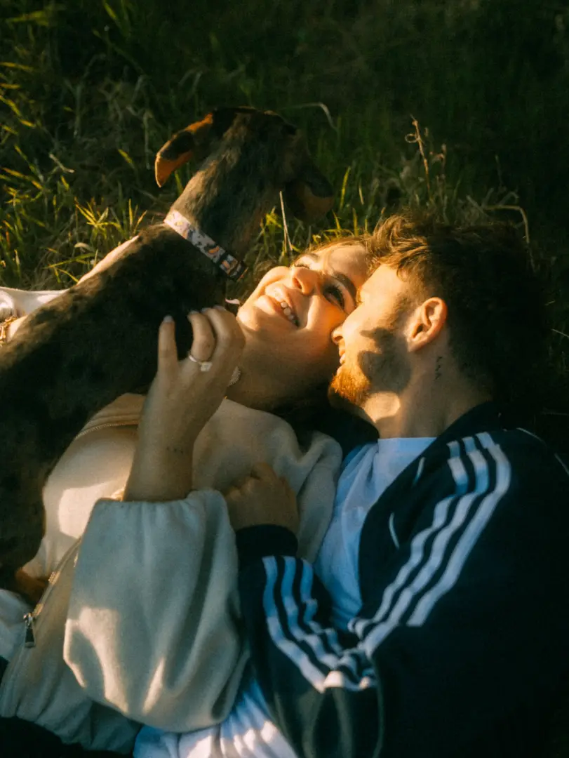 Ein glückliches Paar liegt eng umschlungen im Gras und spielt liebevoll mit seinem Hund, während das warme Licht der Abendsonne für eine romantische Stimmung sorgt. Authentische Pärchenfotos Schwerin, die einen intimen Moment purer Lebensfreude und Verbundenheit einfangen.