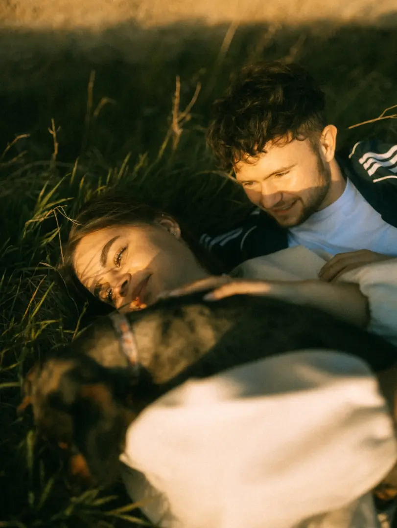 Ein verliebtes Paar genießt bei goldenem Sonnenuntergang entspannte Momente im hohen Gras, während sie gemeinsam liebevoll ihren Hund streicheln. Die warme Lichtstimmung unterstreicht die romantische Atmosphäre dieser Pärchenfotos in Schwerin.