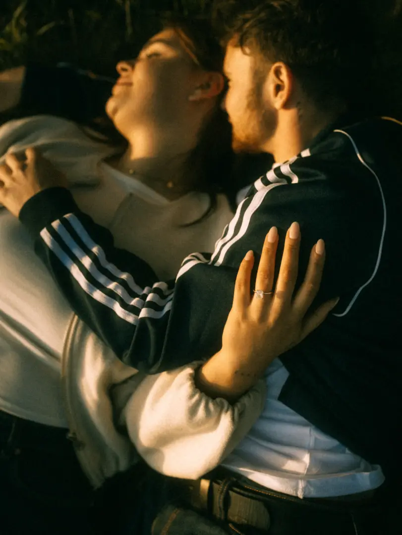 Ein verliebtes Paar liegt eng umschlungen im warmen Licht der Abendsonne im Gras. Die Frau trägt einen Verlobungsring am Finger, während sie ihren Partner liebevoll umarmt. Authentische Pärchenfotos Schwerin, die eine intime und romantische Stimmung einfangen.