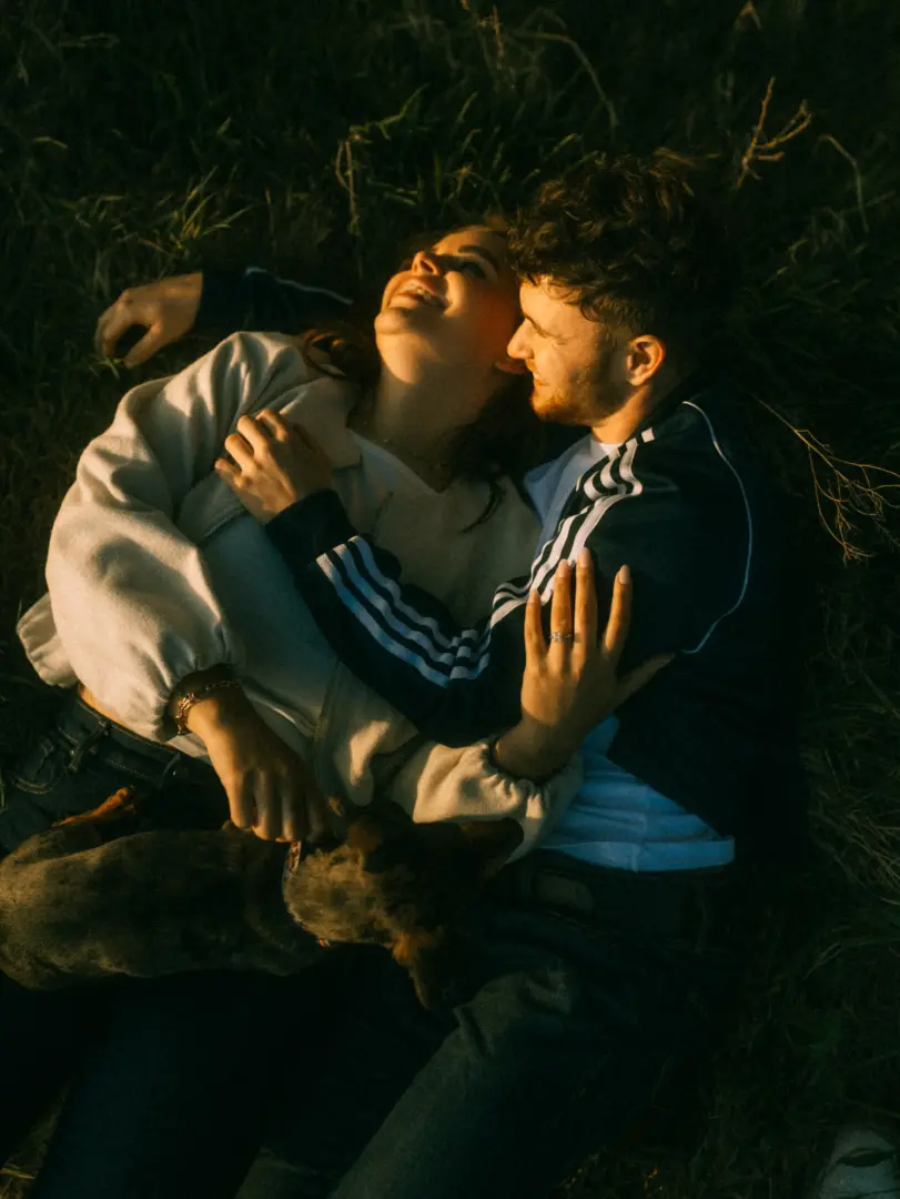 Ein verliebtes Paar lacht herzlich, während es in der goldenen Abendsonne auf einer Wiese liegt und gemeinsam mit seinem Hund kuschelt. Die intime Atmosphäre unterstreicht die Verbundenheit bei diesen authentischen Pärchenfotos Schwerin.