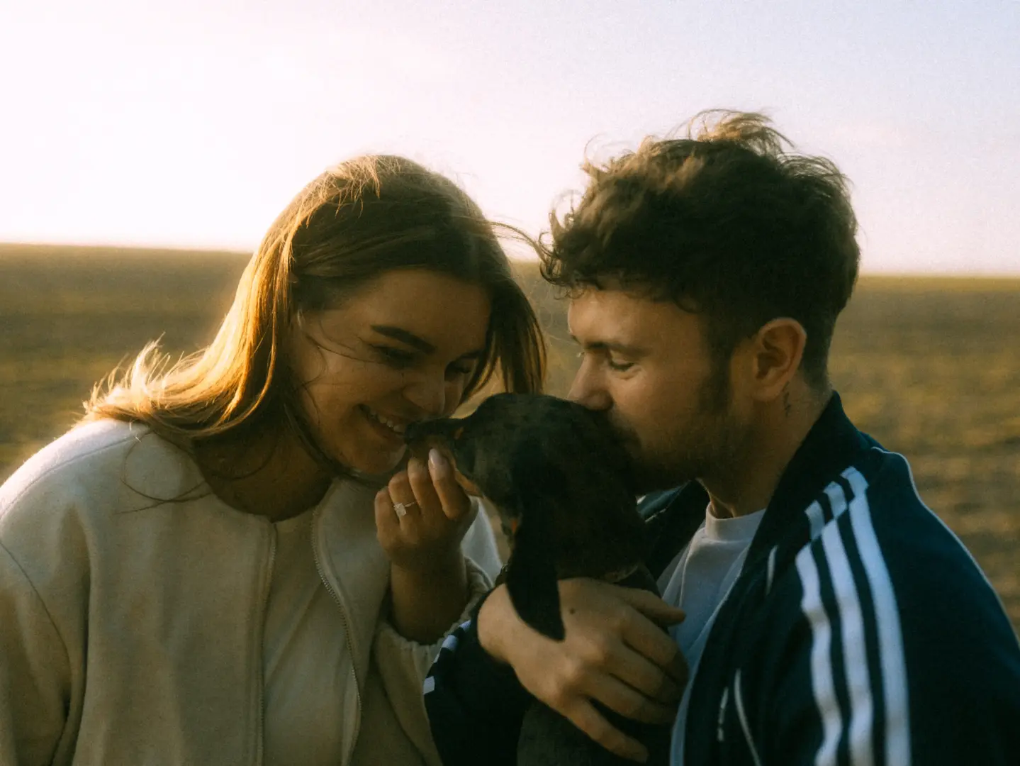 Ein glückliches Paar hält liebevoll seinen kleinen Hund in einem sonnendurchfluteten Feld bei Sonnenuntergang. Die Frau lächelt, während der Mann den Hund küsst, was eine romantische Atmosphäre für Pärchenfotos in Schwerin schafft.