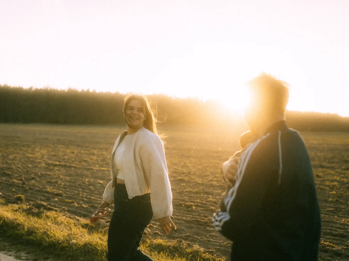 Ein glückliches Paar bei einem Spaziergang während des goldenen Sonnenuntergangs auf einem Feld, festgehalten von einem Fotograf für Pärchen in Schwerin. Die Frau lacht, während sie neben ihrem Partner geht, umgeben von warmem Licht und einer weiten, ländlichen Landschaft.