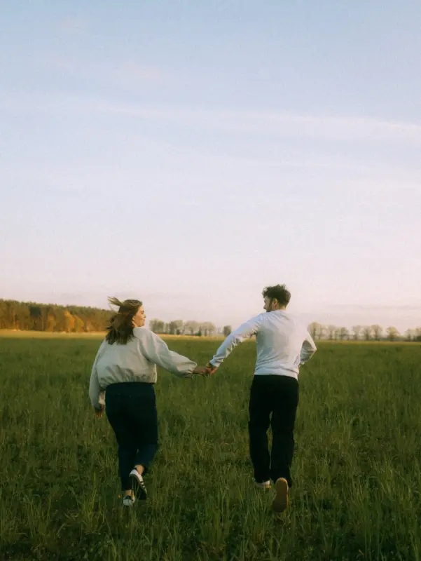 Ein junges Paar läuft händchenhaltend durch eine weite, herbstliche Wiese in der Abendsonne. Die beiden schauen sich lächelnd an, während sie gemeinsam in die Ferne gehen. Authentische Pärchenfotos Schwerin, die eine unbeschwerte und romantische Stimmung einfangen.