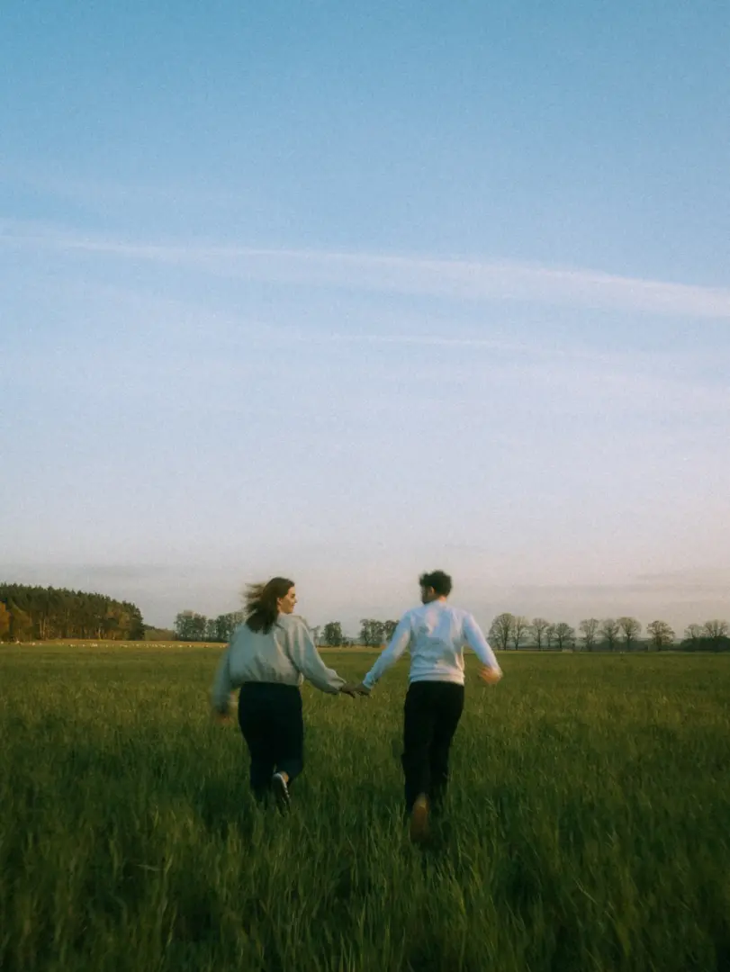 Ein junges Paar läuft Hand in Hand über eine weite, grüne Wiese in der Abenddämmerung. Die beiden blicken sich lächelnd an, während sie durch das hohe Gras rennen. Diese authentischen Pärchenfotos in Schwerin fangen einen romantischen Moment in der Natur ein.