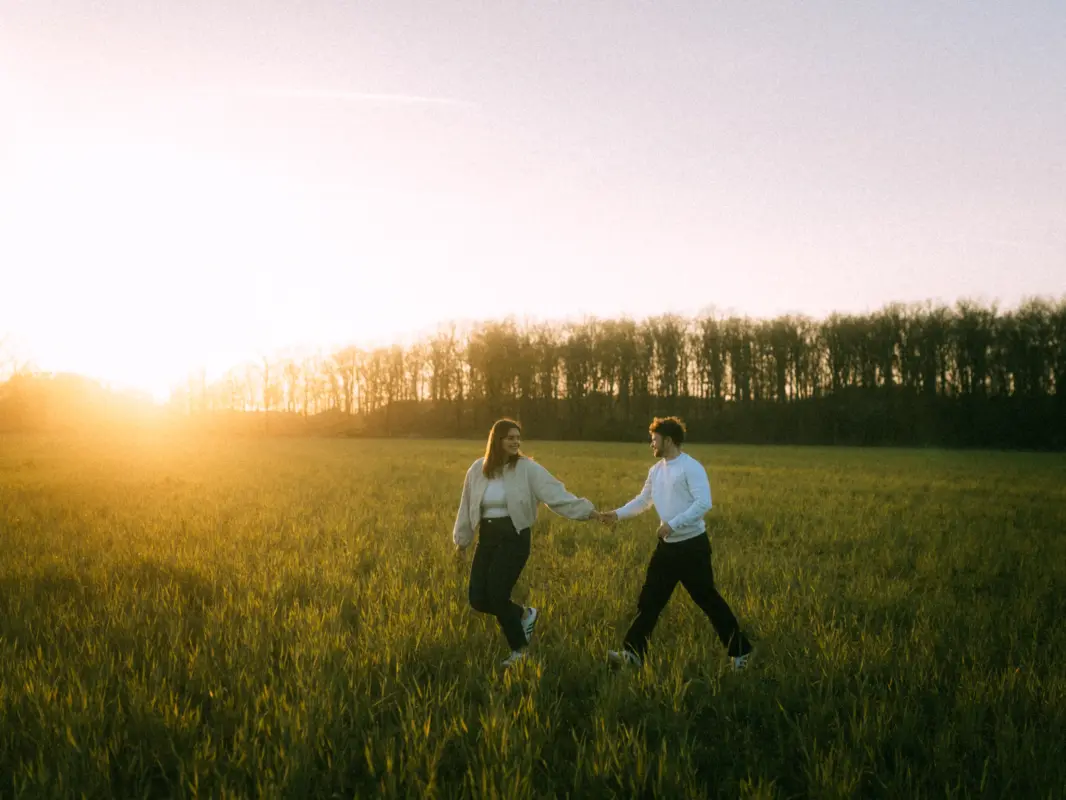 Ein glückliches Paar hält Händchen und läuft bei goldenem Sonnenuntergang durch ein weites, grünes Feld vor einer Waldkulisse. Die entspannte Stimmung und das warme Licht unterstreichen diese authentischen Pärchenfotos in Schwerin.