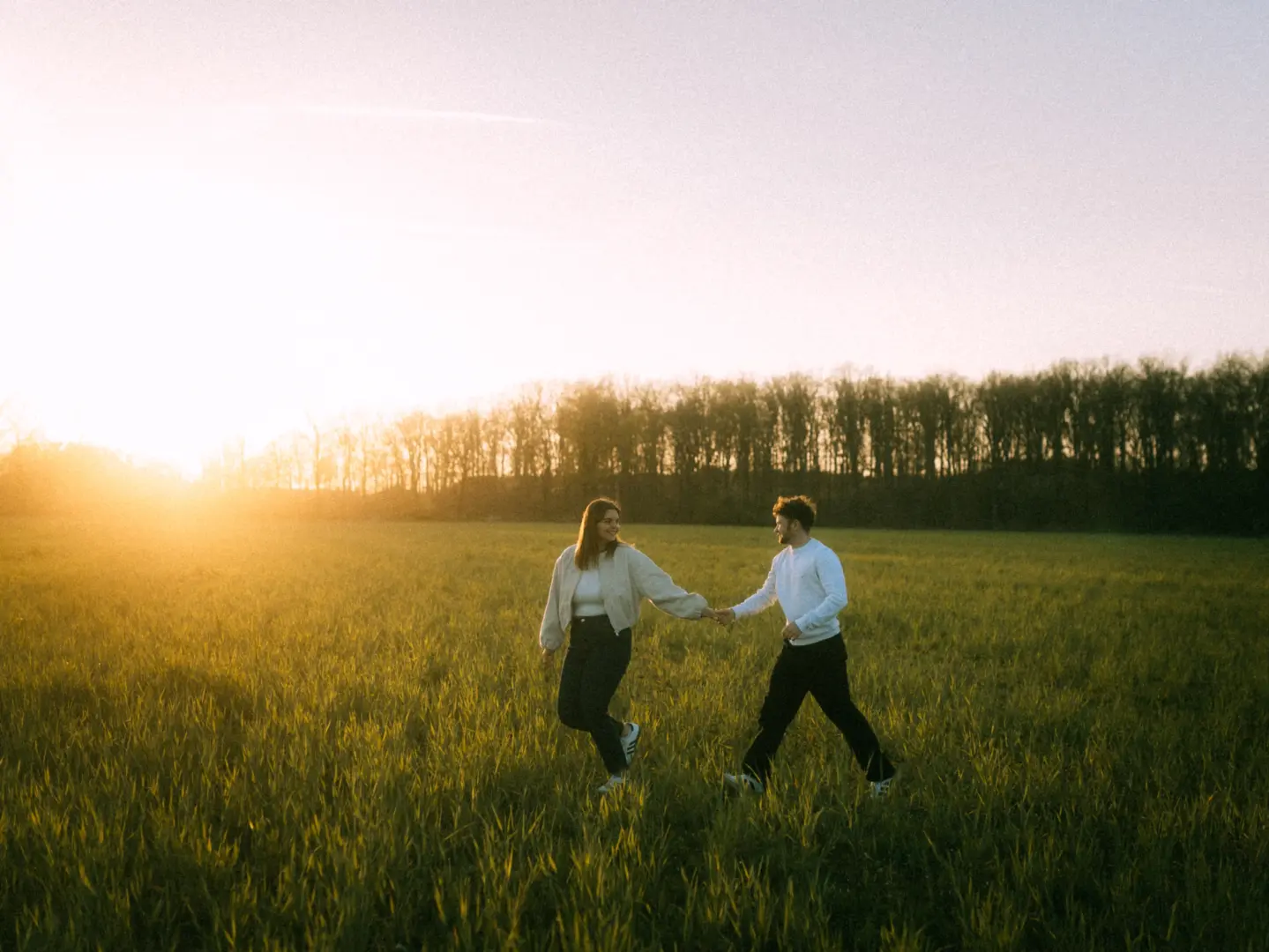 Ein glückliches Paar hält Händchen und läuft bei goldenem Sonnenuntergang durch ein weites, grünes Feld vor einer Waldkulisse. Die entspannte Stimmung und das warme Licht unterstreichen diese authentischen Pärchenfotos in Schwerin.