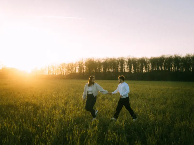 Ein glückliches Paar hält Händchen und läuft bei goldenem Sonnenuntergang durch ein weites, grünes Feld vor einer Waldkulisse. Die entspannte Stimmung und das warme Licht unterstreichen diese authentischen Pärchenfotos in Schwerin.
