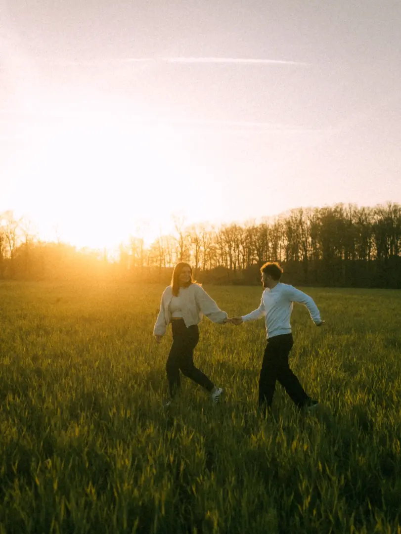 Ein glückliches Paar hält Händchen und läuft lachend durch eine sonnendurchflutete Wiese während des Golden Hour Lichts. Die romantische Stimmung unterstreicht diese authentischen Pärchenfotos Schwerin in der Natur.