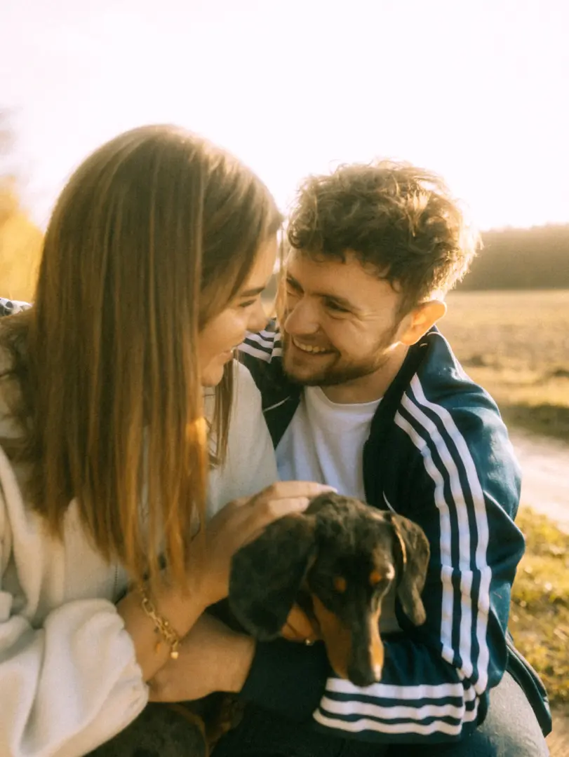 Ein glückliches Paar bei einem Shooting für Pärchenfotos in Schwerin, das gemeinsam einen kleinen Dackel im Arm hält. Die beiden lächeln sich liebevoll an, während sie in einer sonnendurchfluteten, herbstlichen Naturkulisse sitzen.