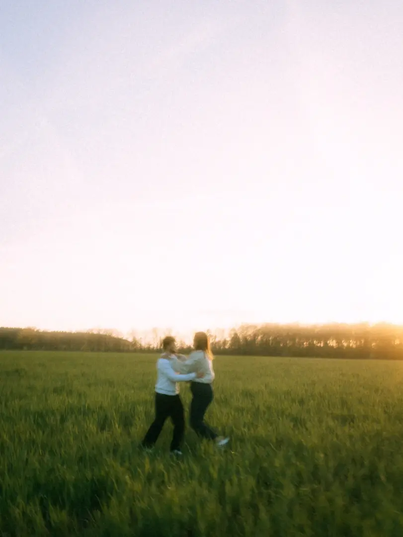 Ein Paar tanzt ausgelassen und glücklich in einem weiten, grünen Feld während des goldenen Sonnenuntergangs. Die beiden tragen helle Oberteile und wirken sehr vertraut bei diesen authentischen Pärchenfotos in Schwerin.