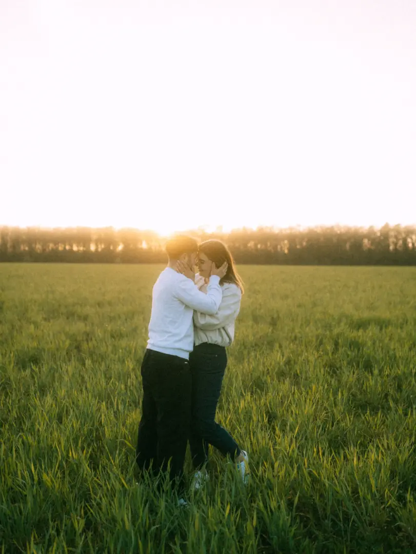 Ein verliebtes Paar steht sich in einem grünen Feld bei Sonnenuntergang gegenüber und hält sich zärtlich an den Wangen, während das warme Gegenlicht eine romantische Stimmung erzeugt. Professionelle Pärchenfotos in Schwerin für unvergessliche Momente.