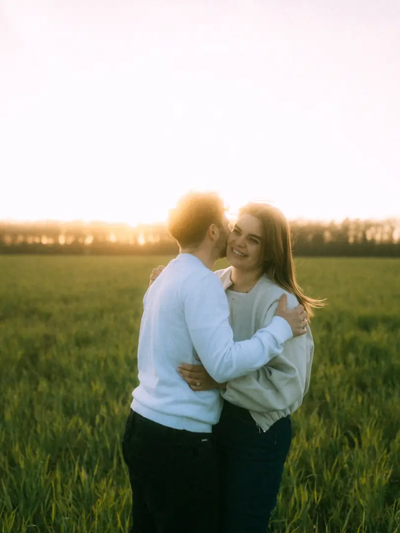 Ein glückliches Paar bei einem romantischen Fotoshooting auf einem Feld im warmen Licht des Sonnenuntergangs, während er ihr einen Kuss auf die Wange gibt. Authentische Pärchenfotos Schwerin, die eine intime und liebevolle Stimmung einfangen.