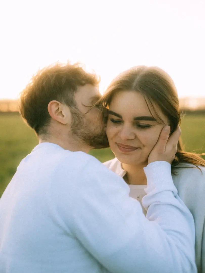 Ein junges Paar bei einem romantischen Fotoshooting im Freien während der goldenen Stunde. Ein Mann mit Bart küsst zärtlich die Wange seiner Partnerin, die ihn sanft an der Wange hält. Authentische Pärchenfotos Schwerin in einer weichen, warmen Lichtstimmung.