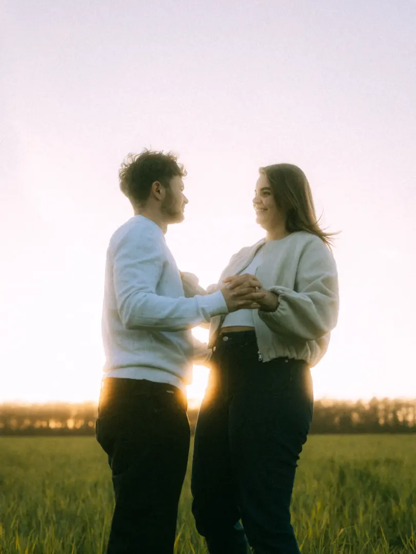 Ein junges Paar hält sich liebevoll an den Händen und lächelt sich glücklich an, während sie bei goldenem Sonnenuntergang in einem weiten Feld stehen. Authentische Pärchenfotos Schwerin, die eine intime und romantische Stimmung einfangen.