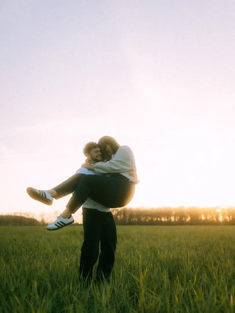 Ein glückliches Paar bei einem romantischen Fotoshooting auf einer grünen Wiese im warmen Licht des Sonnenuntergangs. Der Mann trägt die Frau auf dem Arm, beide lachen ausgelassen. Authentische Pärchenfotos Schwerin für unvergessliche Momente im Freien.