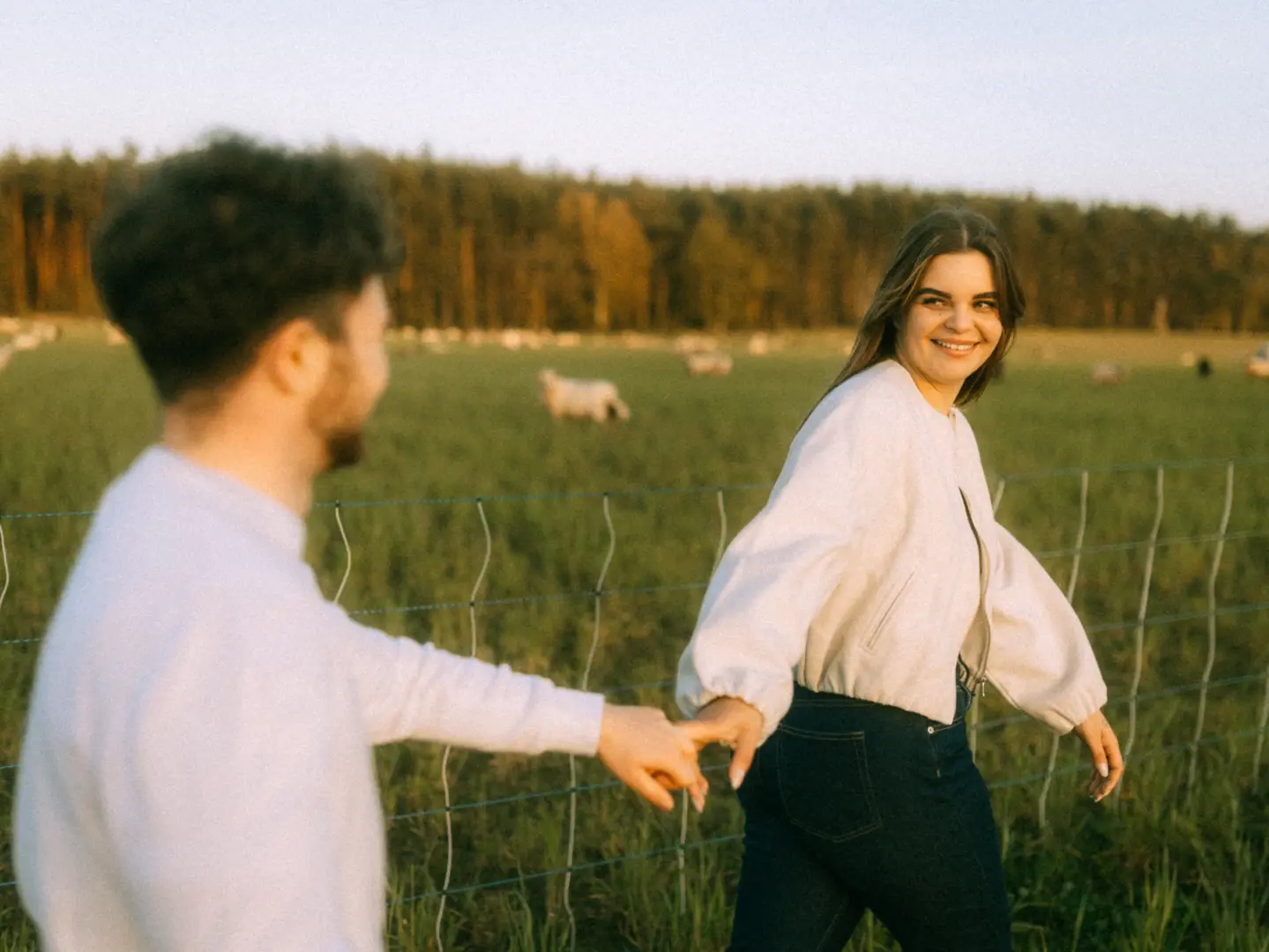 Ein glückliches Paar hält Händchen bei einem Spaziergang auf einer grünen Wiese vor einer Waldkulisse, während die Frau sich lächelnd zu ihrem Begleiter umdreht. Authentische Pärchenfotos Schwerin in natürlicher Abendstimmung.