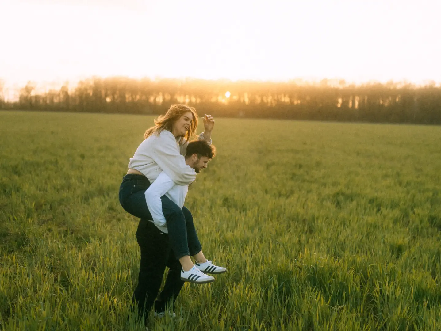 Ein glückliches Paar bei einem spielerischen Huckepack-Ritt auf einer weiten Wiese während des warmen Sonnenuntergangs. Die romantische Stimmung fängt einen unbeschwerten Moment in der Natur ein. Professionelle Pärchenfotos Schwerin.