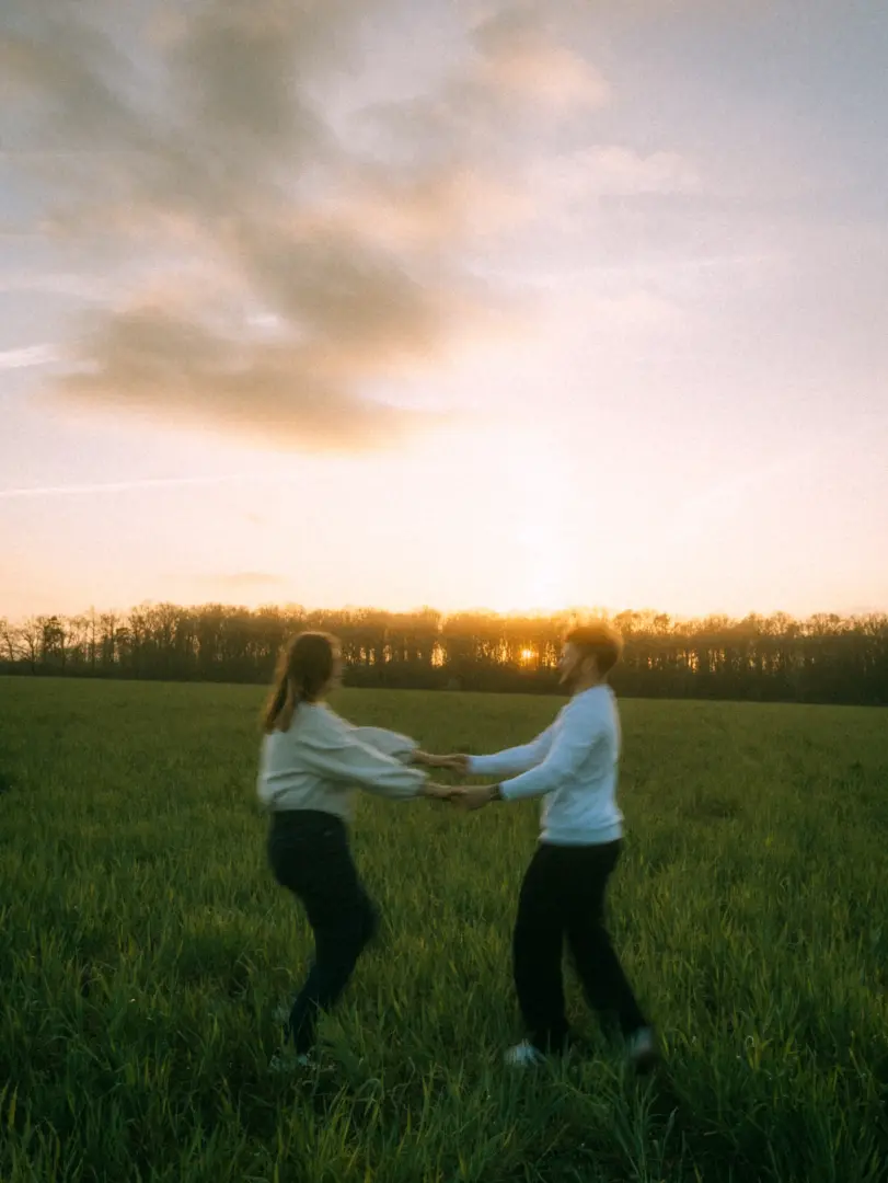 Ein junges Paar hält sich bei Sonnenuntergang auf einem grünen Feld an den Händen und dreht sich glücklich im Kreis. Die warme Abendsonne taucht die Szene in ein goldenes Licht, während im Hintergrund ein Waldrand zu sehen ist – eine romantische Aufnahme für Pärchenfotos in Schwerin.