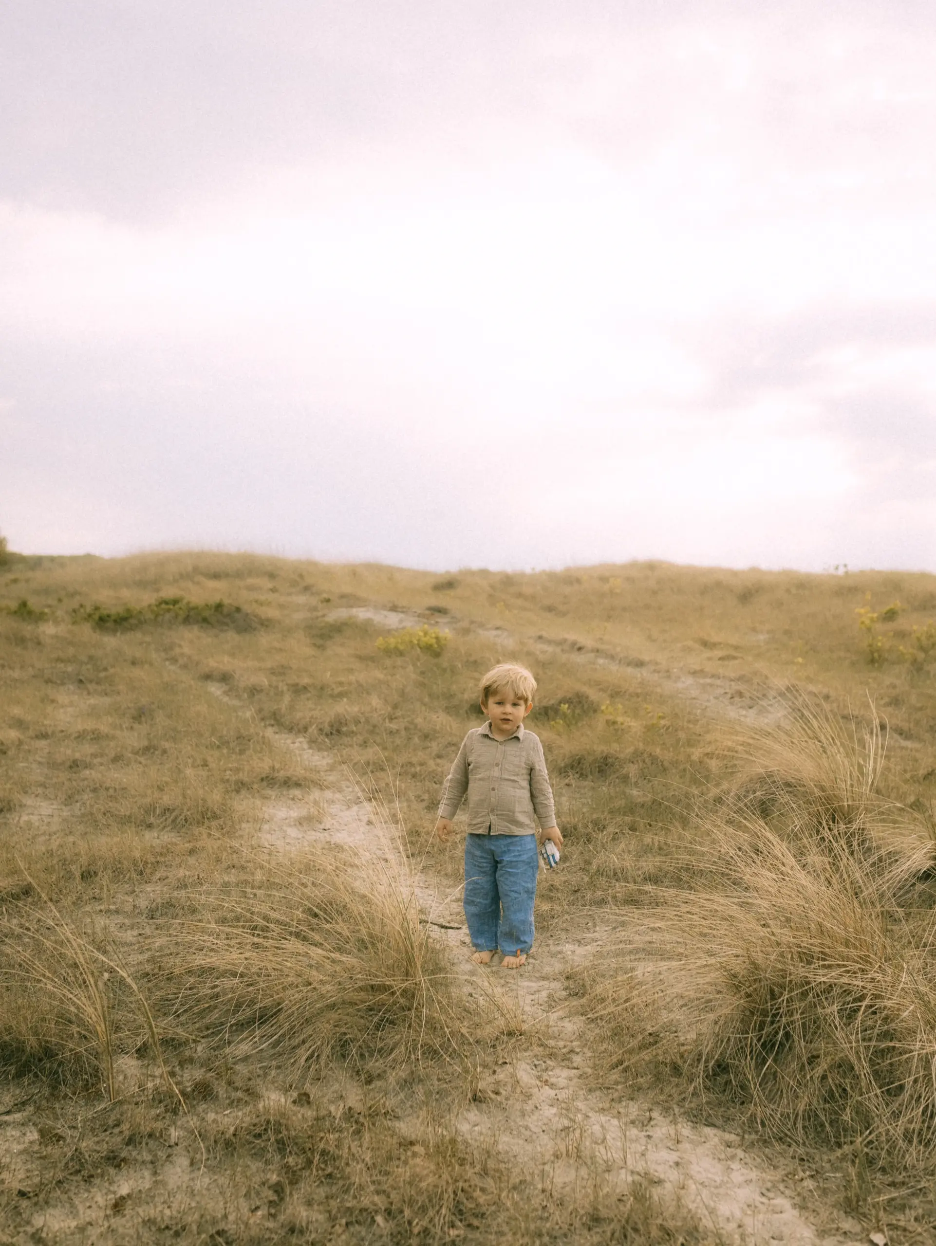 Ein kleiner Junge mit blondem Haar steht barfuß in einer weiten, sonnenverbrannten Dünenlandschaft und hält ein kleines Spielzeug in der Hand. Die natürliche, ruhige Atmosphäre unterstreicht die Arbeit als Familienfotograf in Schwerin.