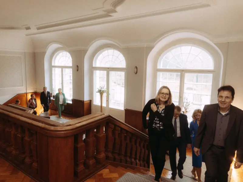 Eine Gruppe von Menschen in eleganter Geschäftskleidung steigt eine prachtvolle, historische Holztreppe in einem hellen Gebäude mit hohen Rundbogenfenstern hinauf. Die Szene vermittelt eine professionelle Atmosphäre, ideal für Business-Porträts oder Veranstaltungen.