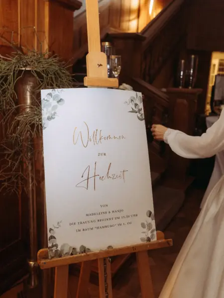Ein elegantes Willkommensschild für die Hochzeit von Madeleine & Hanjo auf einer Holzstaffelei in einem historischen Gebäude. Das Schild mit Eukalyptus-Motiv weist auf die Trauung um 13:30 Uhr im Raum „Hamburg“ hin, aufgenommen von einem Hochzeitsfotograf in Schwerin und MV.