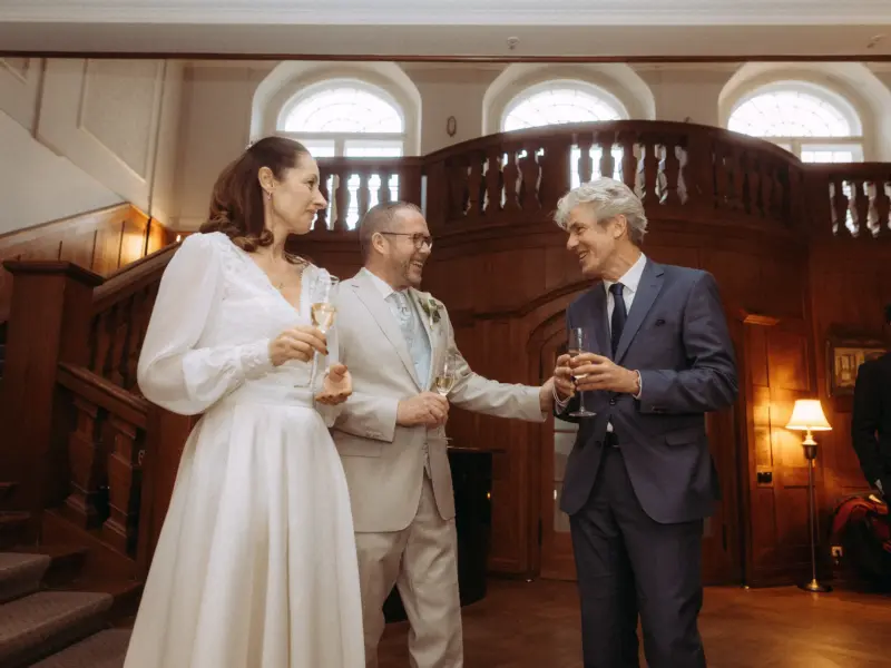 Eine Braut in einem eleganten weißen Hochzeitskleid und ein Bräutigam im hellen Anzug stoßen mit einem Gast in einem eleganten blauen Anzug an. Die festliche Szene findet in einem prunkvollen historischen Saal mit Holztreppe statt. Hochzeitsfotograf in Schwerin und MV.