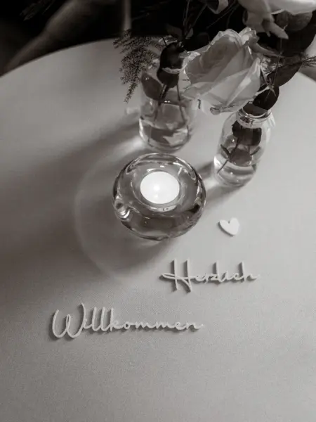 Elegante Tischdekoration für eine Hochzeit mit den Schriftzügen „Herzlich Willkommen“, einer brennenden Kerze in einem Glasgefäß und zwei kleinen Blumenvasen mit Rosen, fotografiert von einem Hochzeitsfotograf in Schwerin und MV.