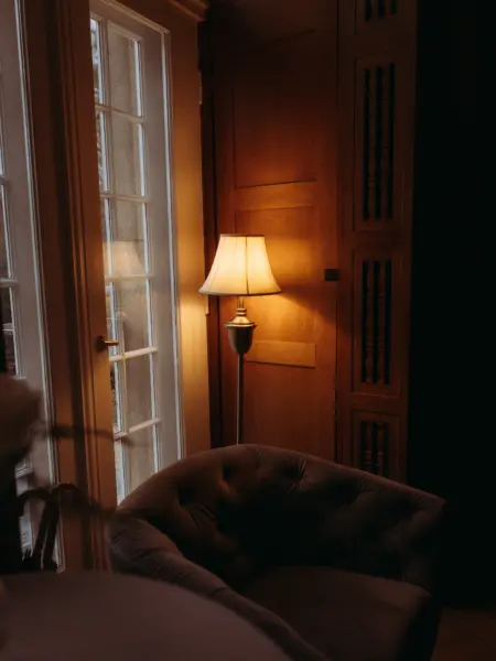 Ein gemütlicher, gepolsterter Sessel steht in einem stimmungsvollen Raum neben einer hohen Fenstertür. Eine klassische Stehlampe sorgt für warmes, sanftes Licht, das die Holzvertäfelung im Hintergrund sanft beleuchtet und eine behagliche Atmosphäre schafft.