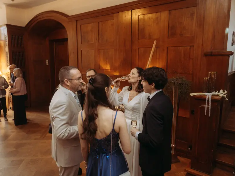 Eine Braut in einem eleganten weißen Brautkleid stößt bei einer Hochzeitsfeier in einem stilvollen Raum mit Holzvertäfelung mit ihren Gästen an. Professionelle Momente eingefangen von Ihrem Hochzeitsfotograf in Schwerin und MV.
