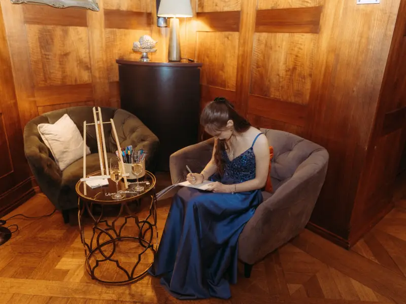 Eine junge Frau in einem eleganten, dunkelblauen Abendkleid sitzt in einem gemütlichen Sessel und schreibt konzentriert in ein Gästebuch. Neben ihr steht ein kleiner Tisch mit Champagnergläsern und einer Staffelei. Hochzeitsfotograf in Schwerin und MV.