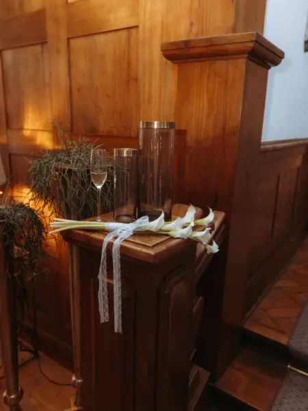 Ein eleganter Brautstrauß aus weißen Calla-Lilien mit zartem Spitzenband liegt auf einem hölzernen Treppenpfosten neben zwei Glasvasen und einem Sektglas – eine stimmungsvolle Aufnahme vom Hochzeitsfotograf in Schwerin und MV.