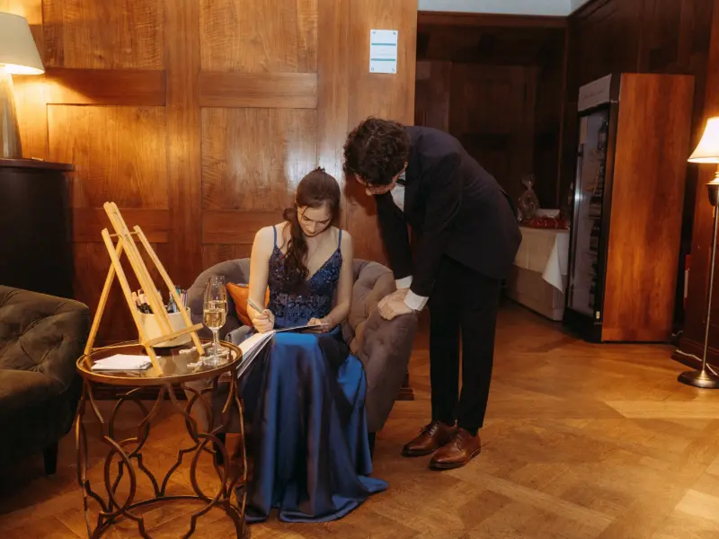 Eine junge Frau in einem eleganten dunkelblauen Abendkleid schreibt in ein Gästebuch, während ein junger Mann in einem dunklen Anzug ihr aufmerksam zuschaut. Die Szene wirkt wie eine Lovestory, festgehalten von einem Lovestory Fotograf in Schwerin und MV in einem stilvollen Raum.