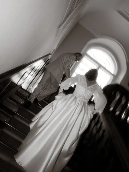Ein Brautpaar in eleganter Hochzeitskleidung geht eine klassische Holztreppe hinauf in Richtung eines hellen, rundbogigen Fensters. Diese romantische Aufnahme wurde von einem Hochzeitsfotograf in Schwerin und MV festgehalten.