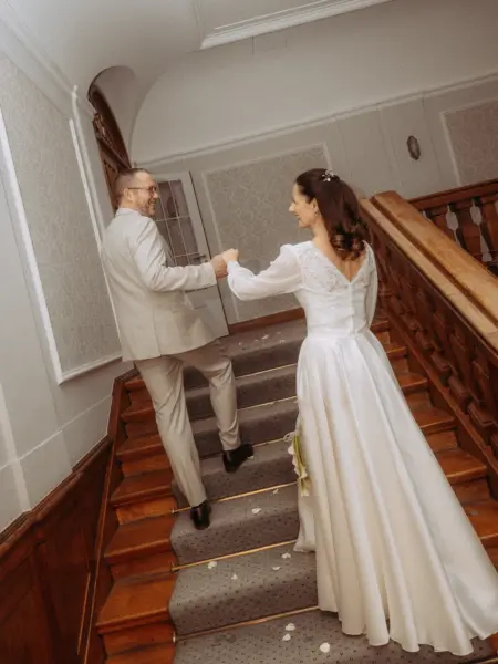 Ein glückliches Brautpaar geht Hand in Hand eine elegante Holztreppe in einem historischen Gebäude hinauf. Die Braut in einem weißen Langarm-Brautkleid blickt lächelnd zu ihrem Bräutigam im hellen Anzug. Eine stimmungsvolle Aufnahme von Ihrem Hochzeitsfotograf in Schwerin und MV.