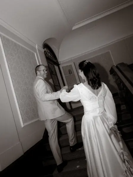 Ein Brautpaar hält Händchen beim Aufstieg einer eleganten Treppe in einem historischen Gebäude. Die Braut trägt ein langärmeliges Brautkleid mit Spitzenbesatz, der Bräutigam einen hellen Anzug. Eine stimmungsvolle Aufnahme vom Hochzeitsfotograf in Schwerin und MV.