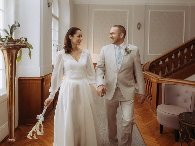 Ein glückliches Brautpaar hält Händchen und lächelt sich in einem eleganten historischen Gebäude mit Holztreppe an. Die Braut trägt ein weißes Langarmkleid mit Blumenstrauß, der Bräutigam einen hellen Anzug. Eine stimmungsvolle Aufnahme vom Hochzeitsfotograf in Schwerin und MV.