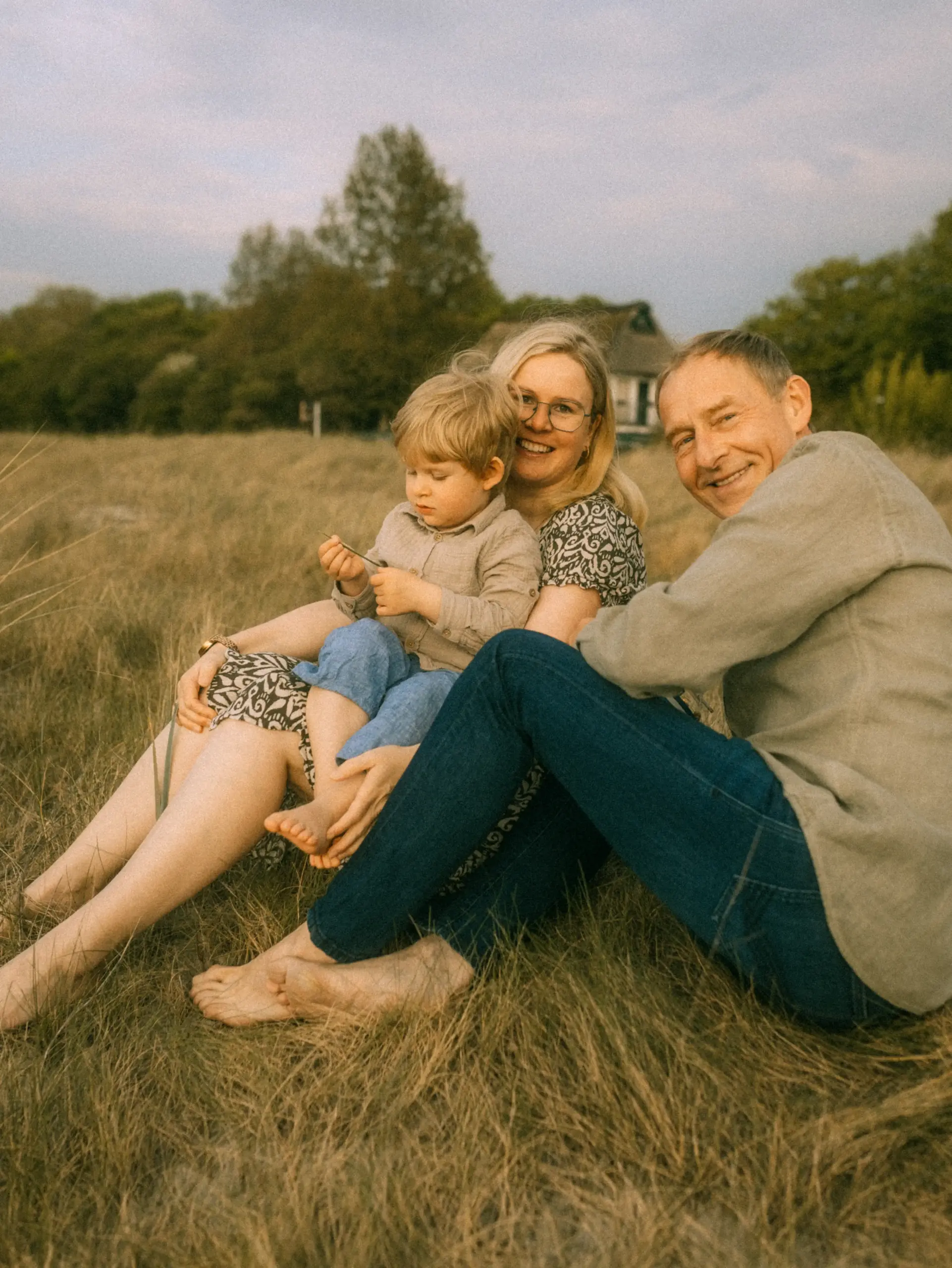 Eine glückliche Familie sitzt barfuß in einer herbstlichen Wiese bei warmem Abendlicht. Mutter, Vater und ihr kleiner Sohn strahlen in die Kamera. Authentische Familienfotos in Schwerin, die eine liebevolle Verbindung in der Natur einfangen.
