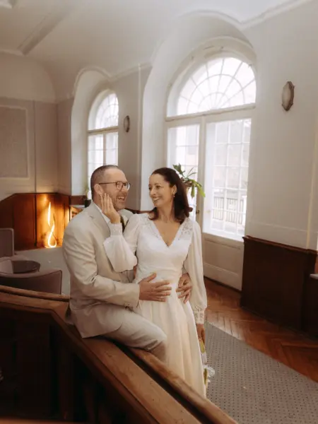 Ein glückliches Brautpaar bei einer intimen Hochzeit: Der Bräutigam im hellen Anzug sitzt auf einem Holzgeländer, während die Braut in einem eleganten weißen Kleid mit langen Ärmeln liebevoll seine Wange berührt. Hochzeitsfotograf in Schwerin und MV.