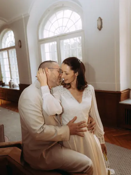 Ein frisch vermähltes Paar in einem hellen, eleganten Saal mit großen Bogenfenstern. Die Braut im weißen Kleid und der Bräutigam im beigen Anzug halten sich liebevoll im Arm und schauen sich tief in die Augen. Authentische Lovestory Fotograf in Schwerin und MV.