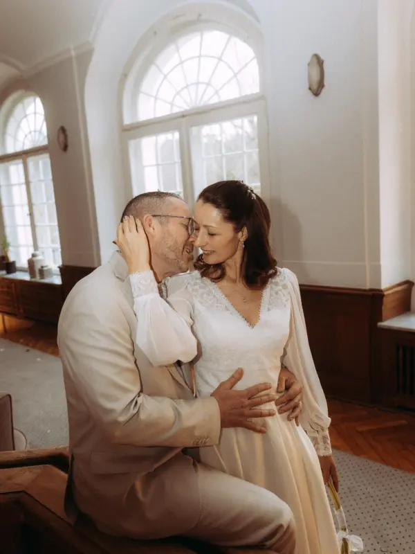 Ein frisch vermähltes Paar in einem hellen, eleganten Saal mit großen Bogenfenstern. Die Braut im weißen Kleid und der Bräutigam im beigen Anzug halten sich liebevoll im Arm und schauen sich tief in die Augen. Authentische Lovestory Fotograf in Schwerin und MV.