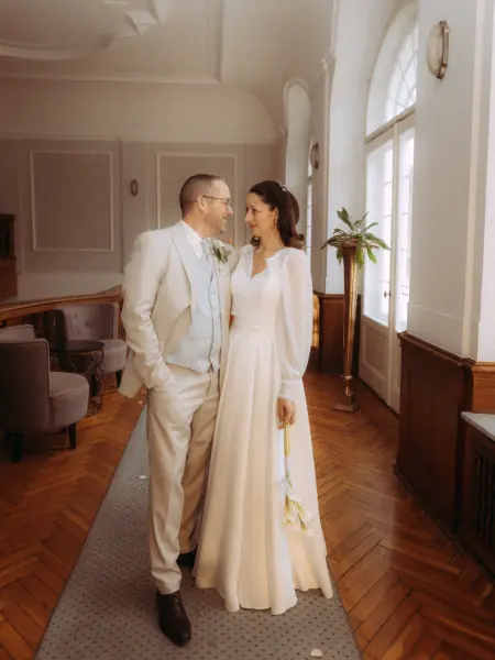 Ein Brautpaar blickt sich liebevoll in einem eleganten, lichtdurchfluteten Flur an. Die Braut trägt ein weißes Kleid mit langen Ärmeln und hält Callas, der Bräutigam einen hellen Anzug. Eine stimmungsvolle Aufnahme von einem Hochzeitsfotograf in Schwerin und MV.