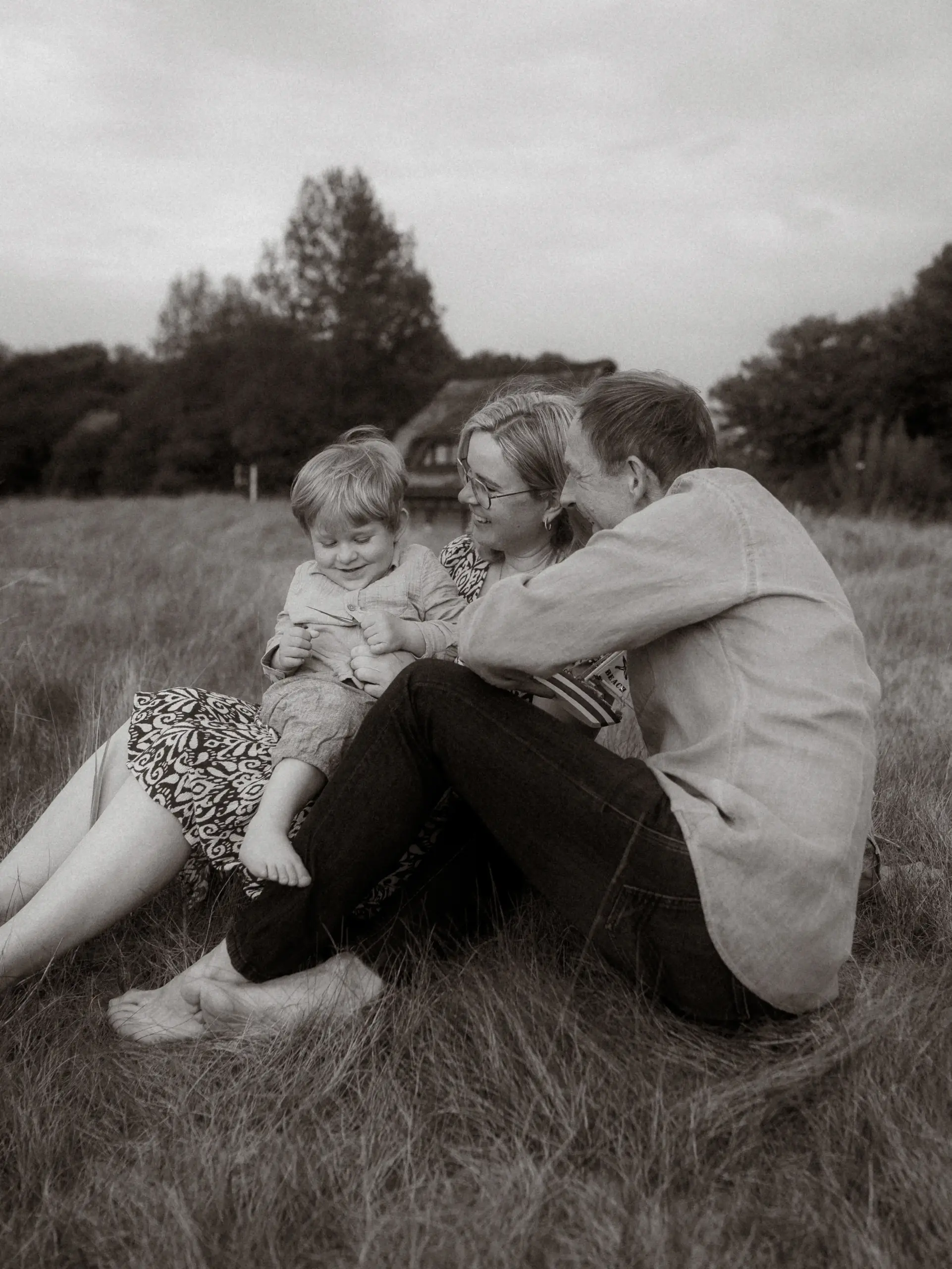 Eine junge Familie genießt einen entspannten Moment auf einer Wiese. Die Eltern sitzen im Gras und lachen gemeinsam mit ihrem kleinen Sohn, der auf dem Schoß der Mutter spielt. Diese authentischen Familienfotos in Schwerin halten die liebevolle Verbindung der drei fest.