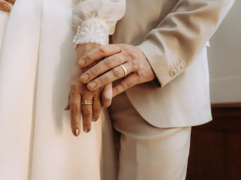 Nahaufnahme eines Brautpaares, bei dem der Bräutigam sanft die Hand der Braut hält. Beide tragen ihre goldenen Eheringe, während die Braut ein weißes Kleid mit Spitzenärmeln und der Bräutigam einen hellen Anzug trägt – ein romantischer Moment eingefangen vom Hochzeitsfotograf in Schwerin und MV.