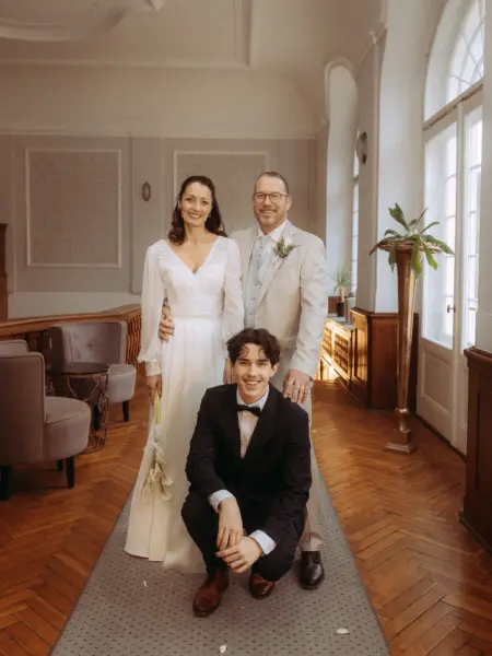 Ein glückliches Brautpaar steht in einem eleganten Saal, während ein junger Mann im Anzug davor kniet. Die Braut trägt ein weißes Spitzenkleid, der Bräutigam einen hellen Anzug. Diese Aufnahme zeigt die Arbeit als Hochzeitsfotograf in Schwerin und MV in einem stilvollen Ambiente.