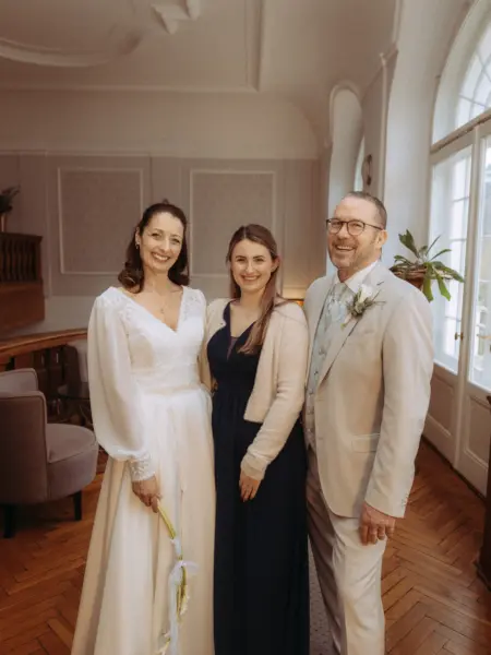 Ein glückliches Brautpaar posiert mit einer jungen Frau in einem eleganten, lichtdurchfluteten Saal. Die Braut trägt ein weißes Hochzeitskleid mit langen Ärmeln, der Bräutigam einen beigen Anzug. Eine schöne Erinnerung festgehalten durch Ihren Hochzeitsfotograf in Schwerin und MV.