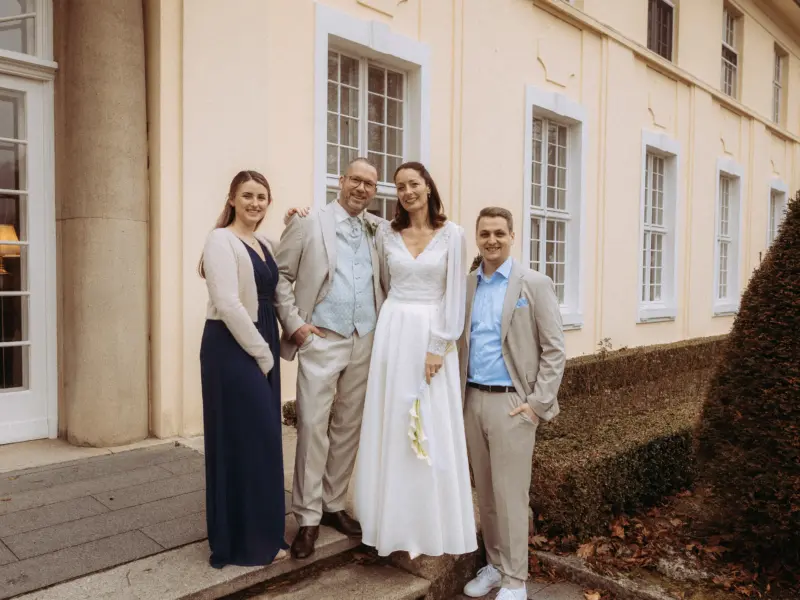 Ein glückliches Brautpaar posiert an seinem Hochzeitstag gemeinsam mit zwei Gästen vor der beigen Fassade eines historischen Gebäudes. Die Braut trägt ein elegantes weißes Brautkleid, der Bräutigam einen hellen Anzug. Hochzeitsfotograf in Schwerin und MV.