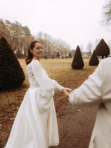 Eine strahlende Braut in einem eleganten weißen Kleid mit langen Ärmeln hält lächelnd die Hand ihres Partners, während sie durch einen herbstlichen Park mit kegelförmigen Bäumen spaziert. Ein romantischer Moment, eingefangen von einem Hochzeitsfotograf in Schwerin und MV.