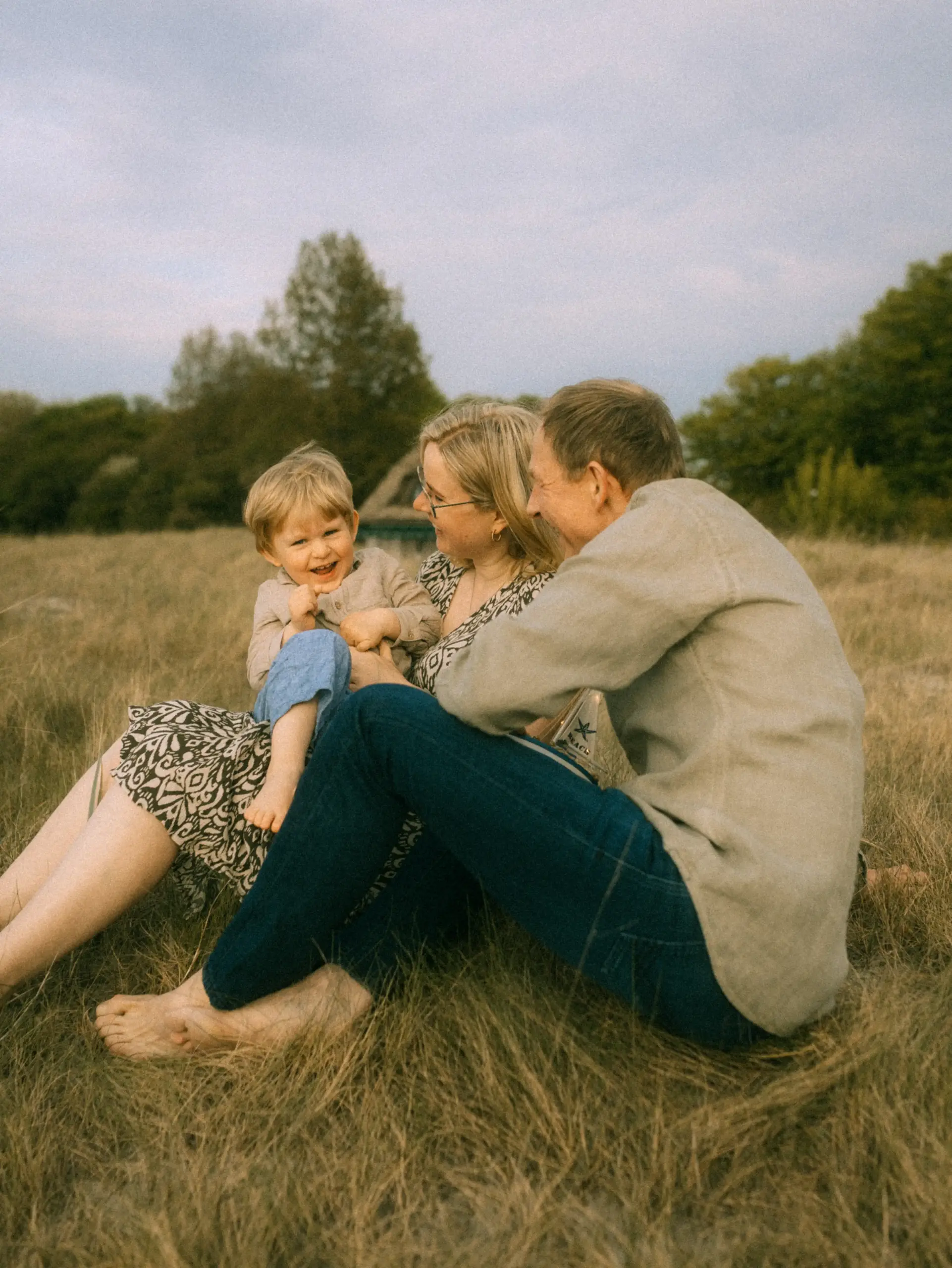 Ein glückliches Paar mit ihrem kleinen Sohn beim Spielen auf einer Wiese im goldenen Abendlicht. Die Familie sitzt entspannt im hohen Gras, während das Kind herzlich lacht – ein authentischer Moment, festgehalten von einem Fotograf für Familien in Schwerin.
