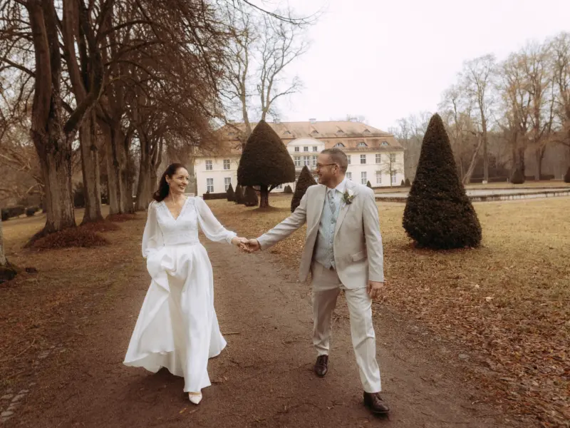 Ein glückliches Brautpaar hält Händchen und spaziert lachend auf einem Kiesweg vor einem historischen Schloss. Die Braut trägt ein elegantes weißes Kleid mit langen Ärmeln, der Bräutigam einen hellen Anzug. Hochzeitsfotograf in Schwerin und MV für unvergessliche Momente.
